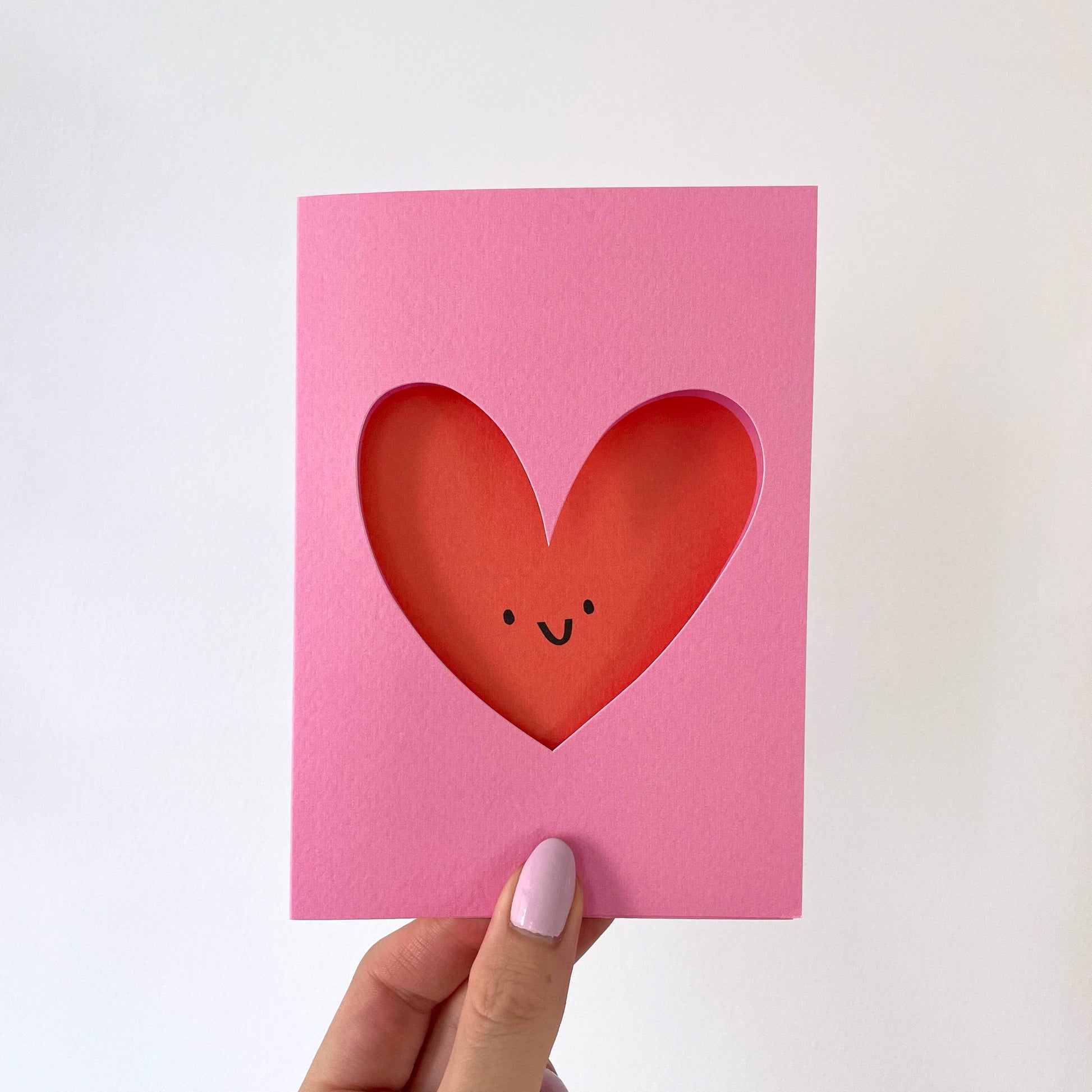 Pink card with a red heart cutout featuring a smiley face, held by a hand on a white background.