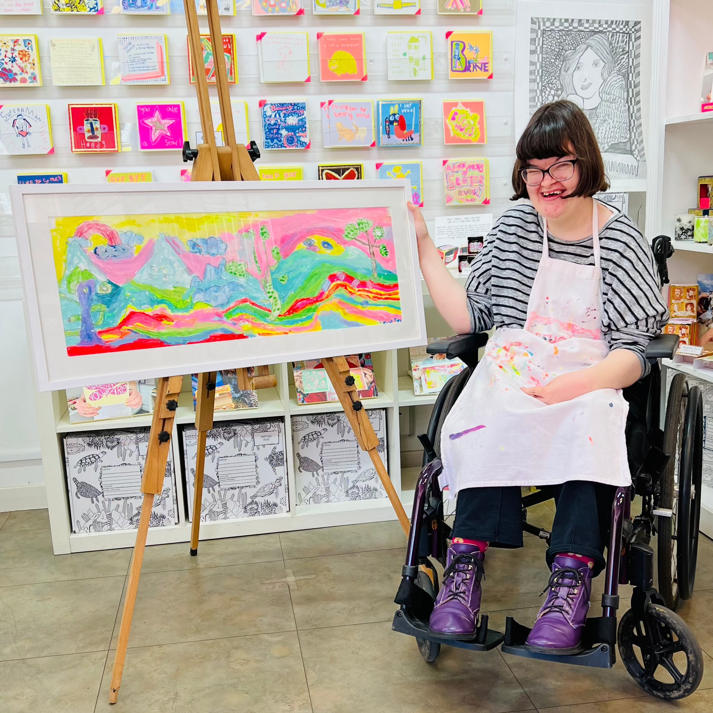 Person in a wheelchair holding a colorful painting in an art studio.