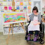 Person in a wheelchair holding a colorful painting in an art studio.