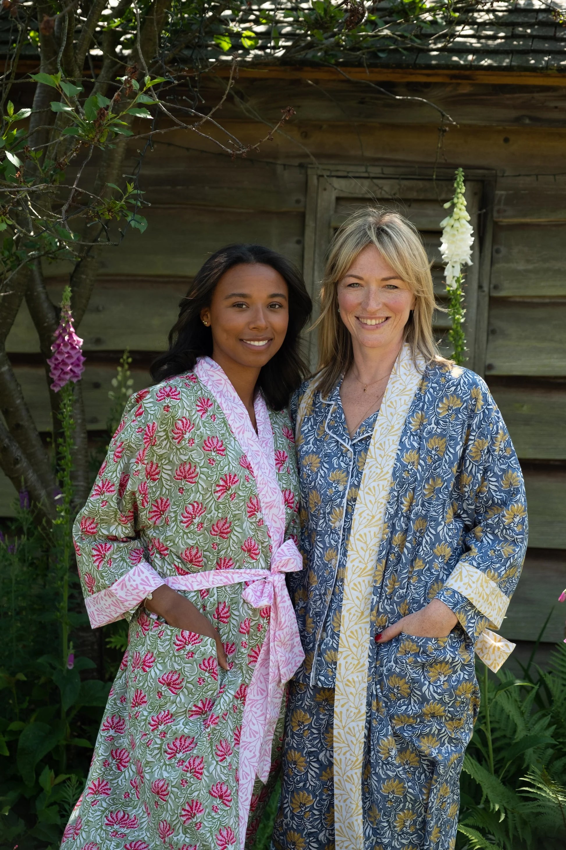 Two women wearing floral robes standing outdoors with a wooden cabin in the background.
