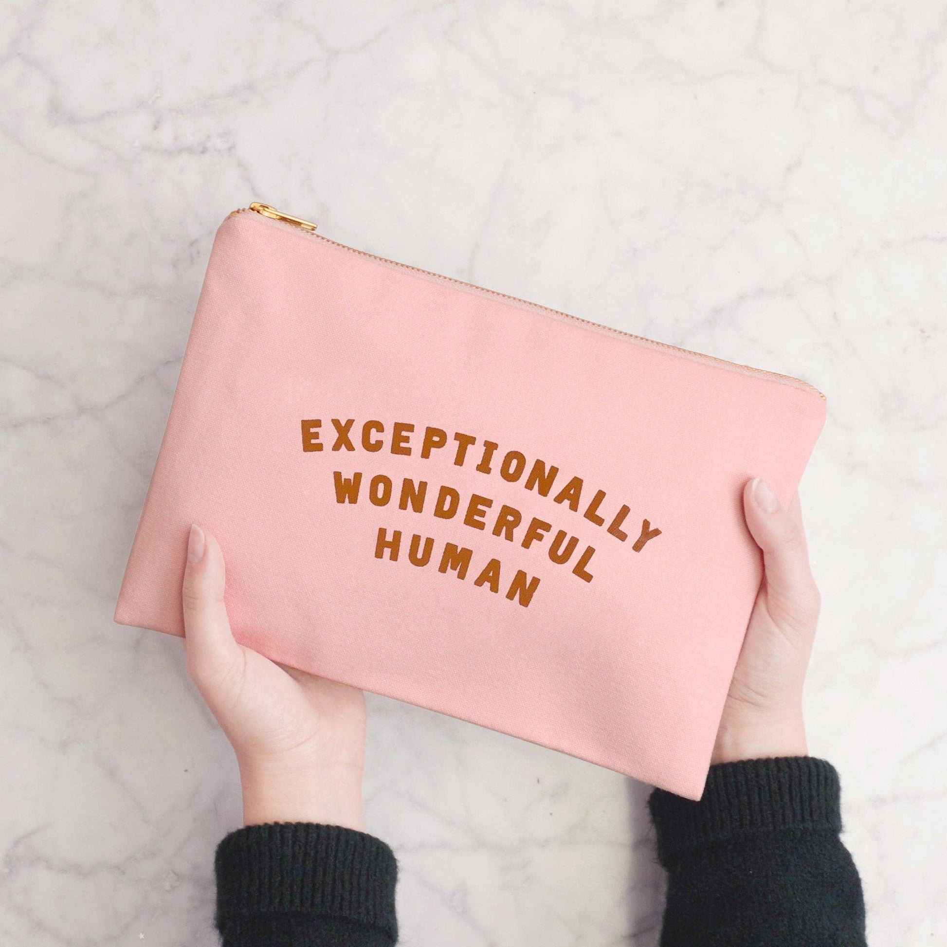 Pink pouch with 'Exceptionally Wonderful Human' text held by a person on a light gray background