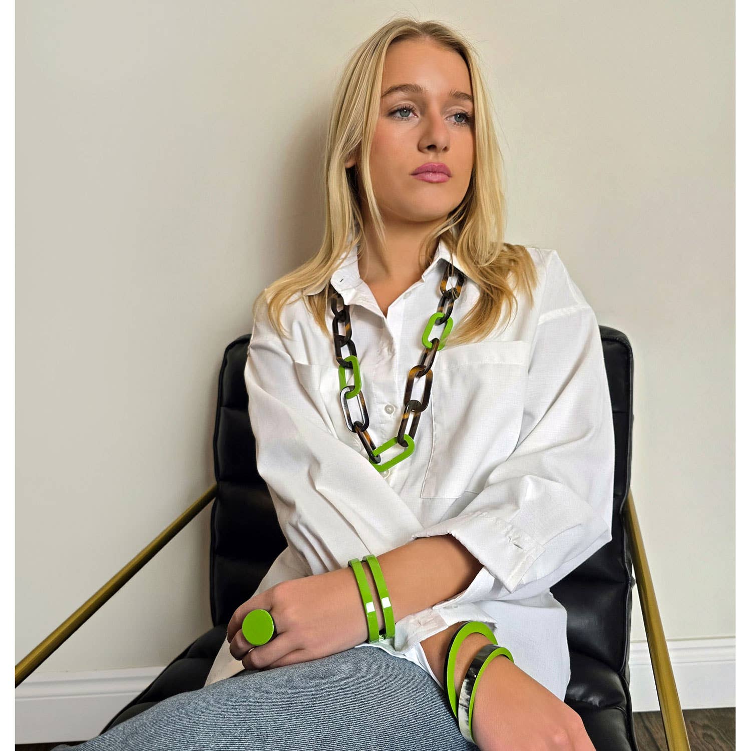 Woman wearing a white shirt with green and black accessories sitting on a chair.