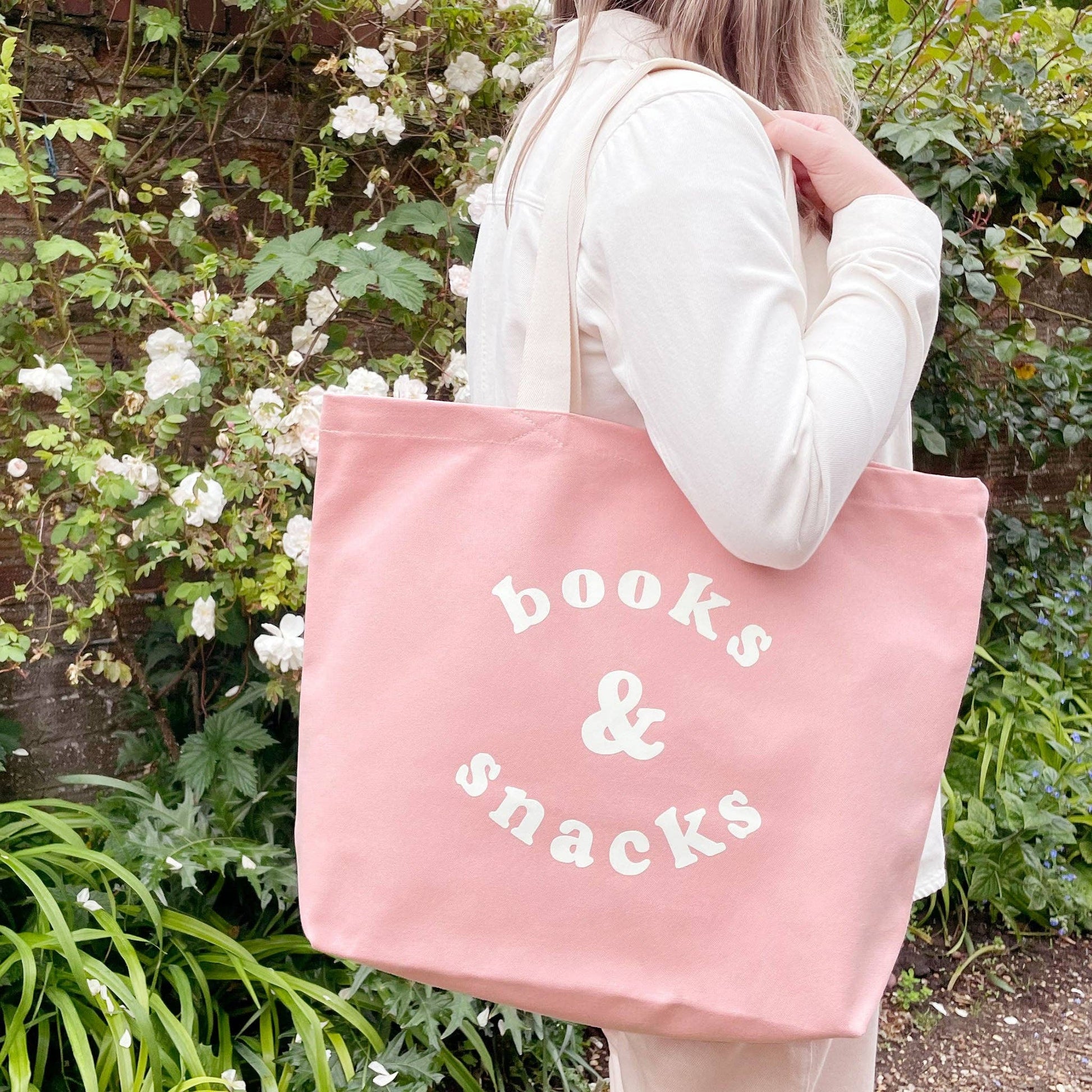 Person holding a pink tote bag with 'books & snacks' text in a garden setting