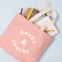 Pink tote bag with 'books & snacks' text, filled with books on a light blue background