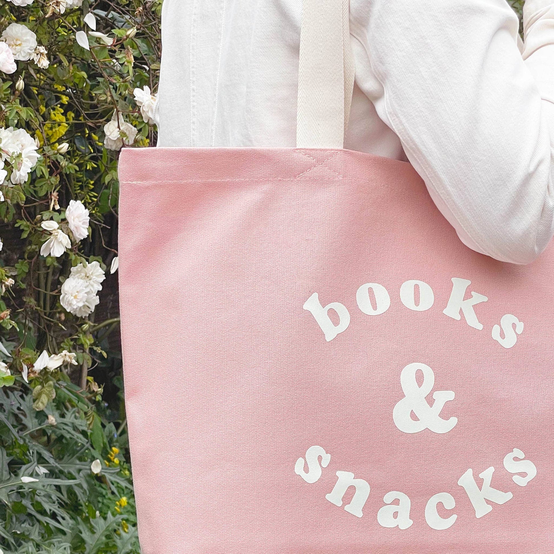 Pink tote bag with 'books & snacks' text held by a person outdoors.