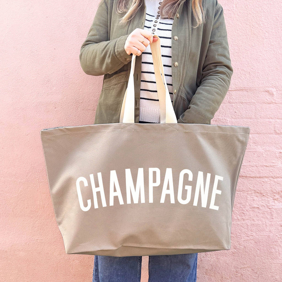 Person holding a beige tote bag with 'CHAMPAGNE' text against a pink background