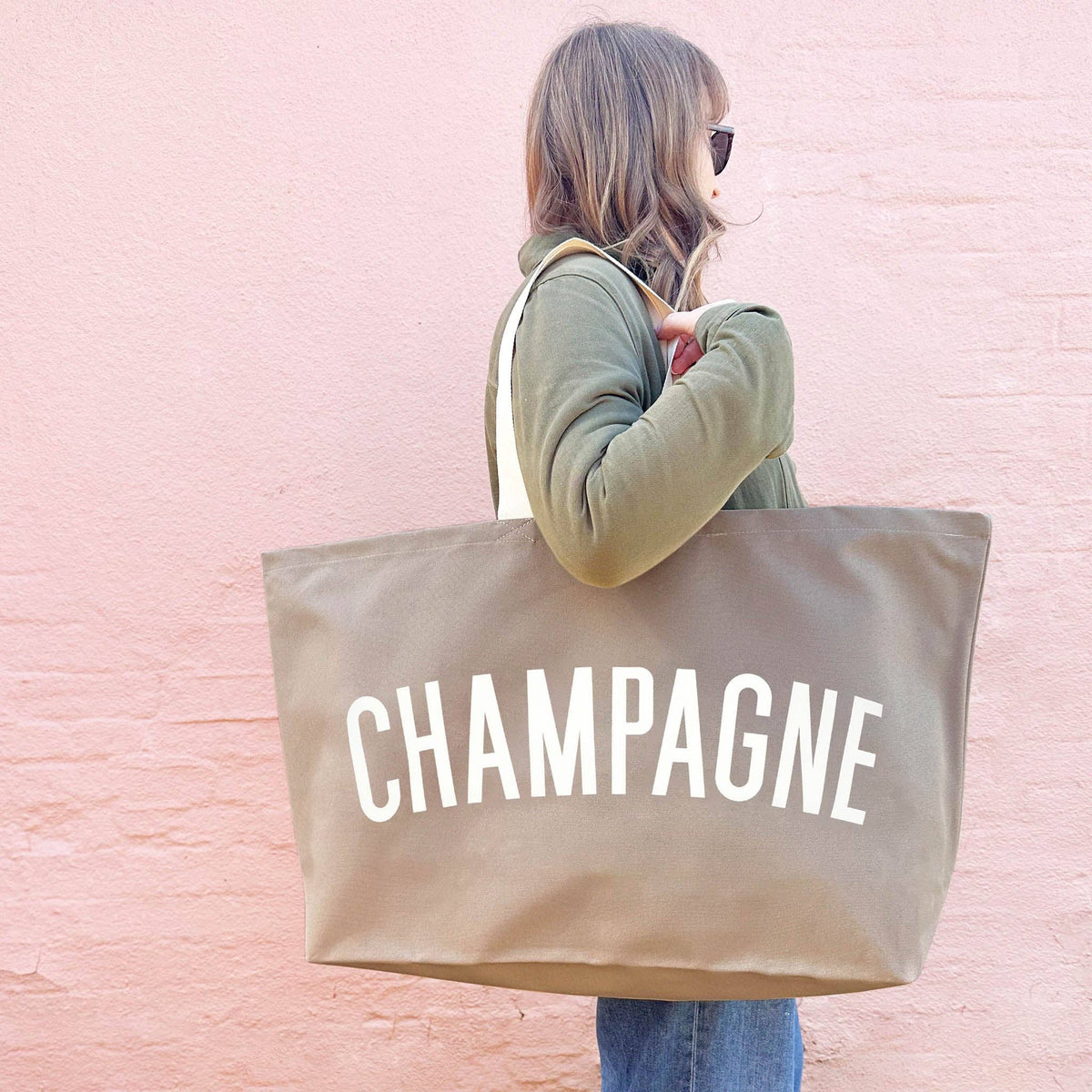 Person holding a beige tote bag with 'CHAMPAGNE' text against a pink brick wall.