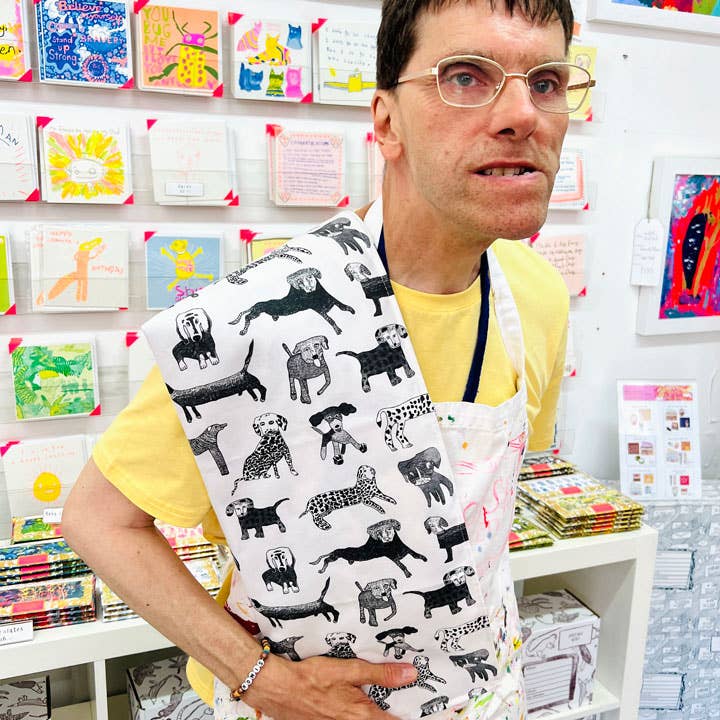 Person wearing a white apron with black dog patterns in front of a display of colorful greeting cards.