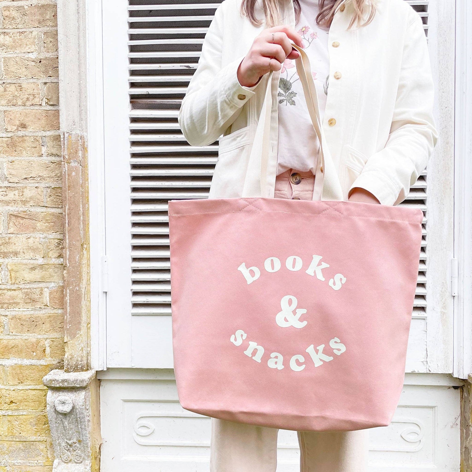Person holding a pink tote bag with 'books & snacks' text against a white shutter background.