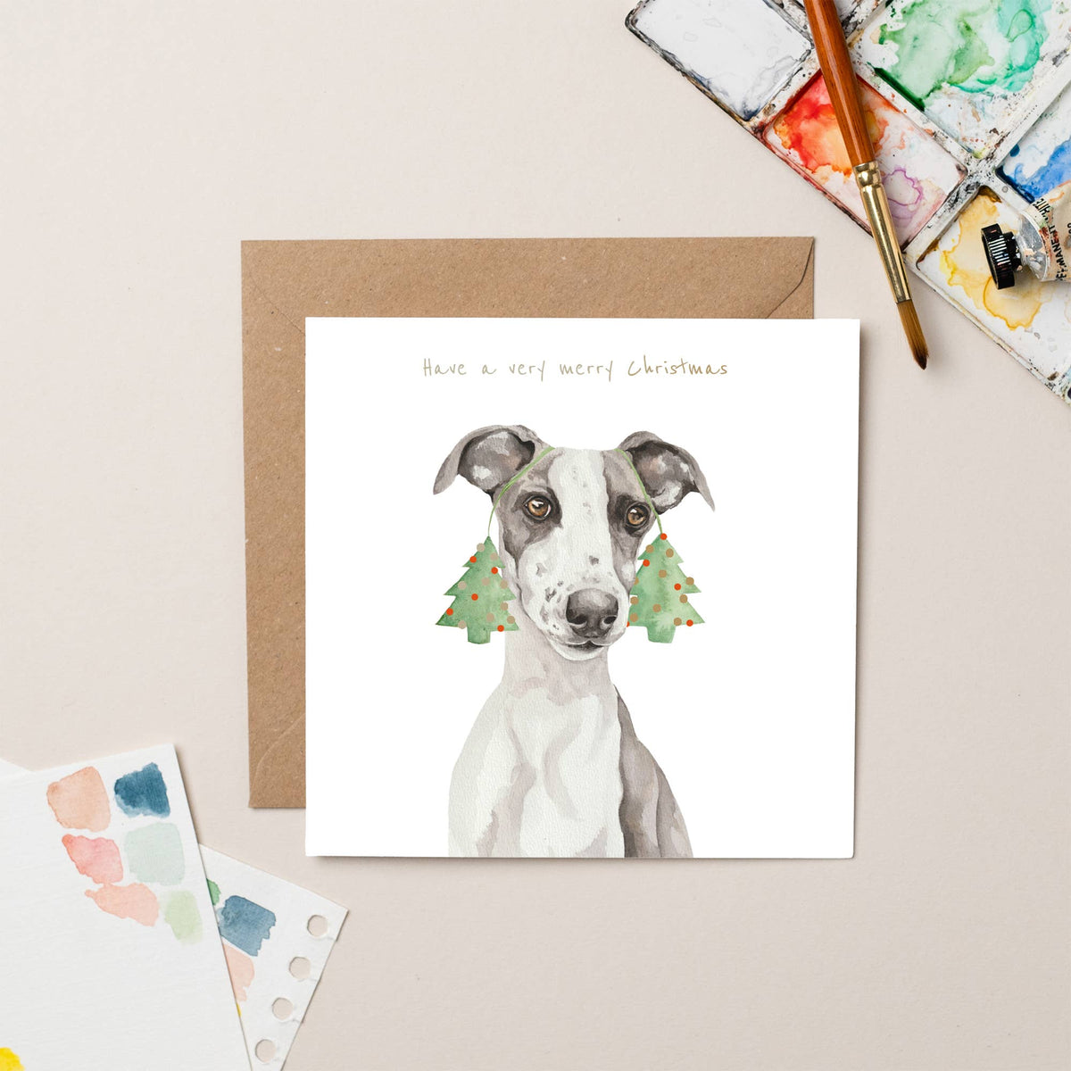 Christmas card with a dog illustration and festive trees, placed on a beige surface with art supplies.