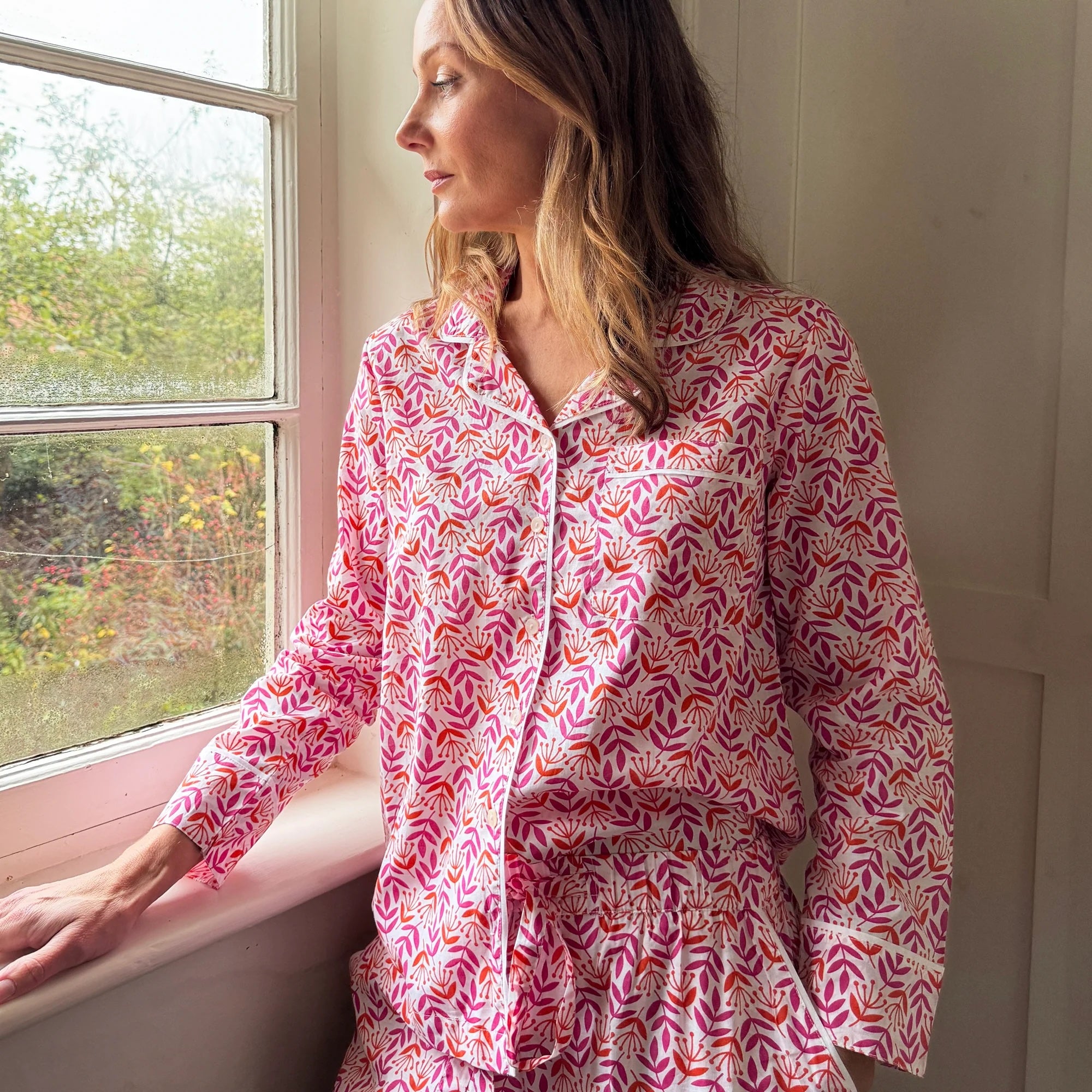 Woman wearing a pink patterned dress sitting by a window.