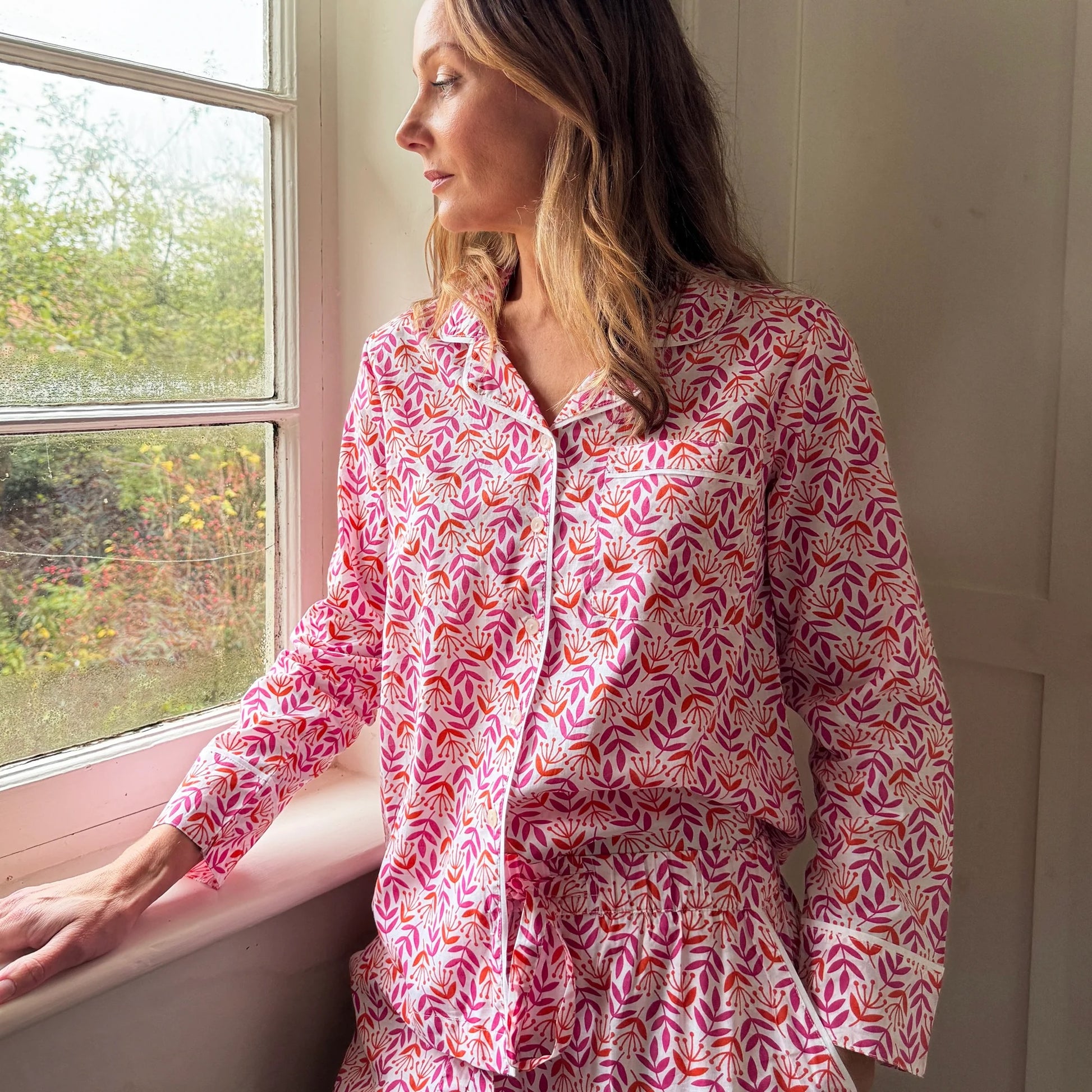 Woman wearing a pink patterned dress sitting by a window.