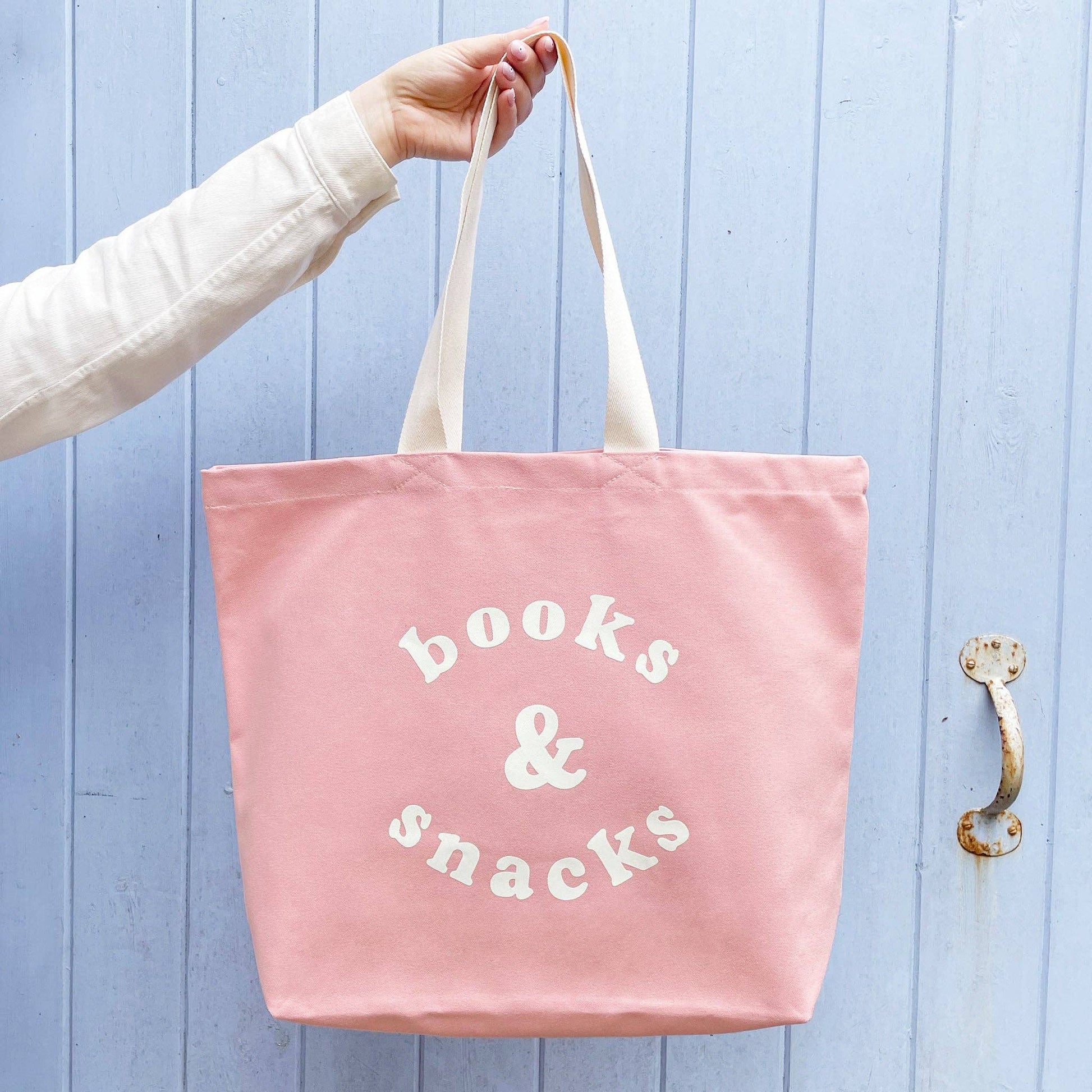 Pink tote bag with 'books & snacks' text held by a person against a light blue wooden door.