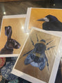 Three artistic prints of a rabbit, raven, and bee on a reflective surface.