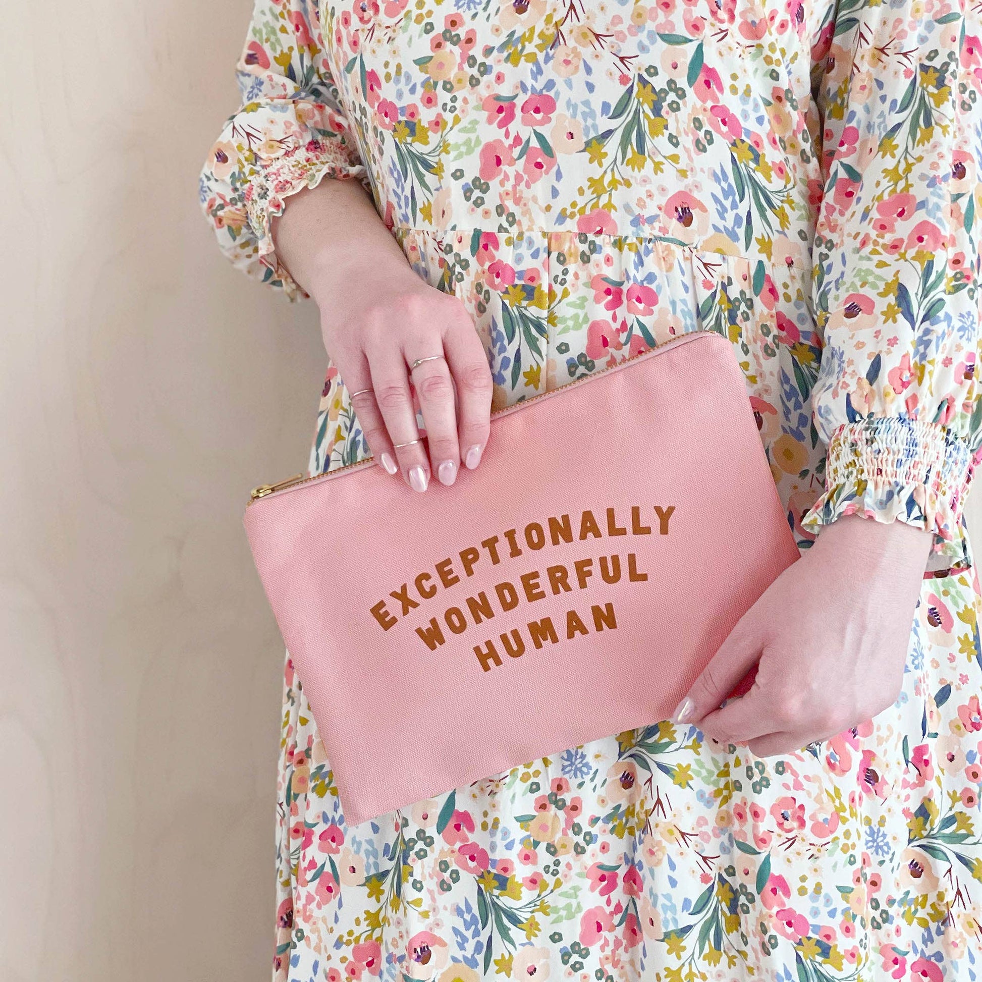 Person holding a pink pouch with 'Exceptionally Wonderful Human' text, wearing a floral dress.