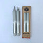 Two gray candles with silver bottoms next to their cardboard packaging on a white background
